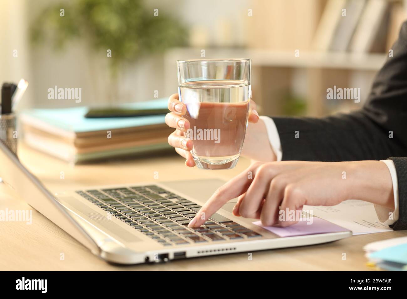 Gros plan de femme d'entrepreneur mains tenant un verre d'eau travaillant sur ordinateur portable à la maison Banque D'Images