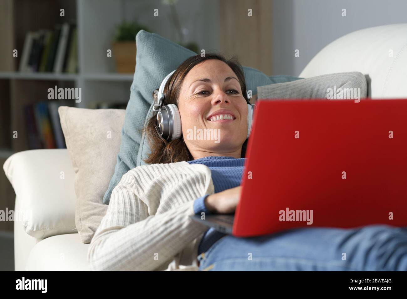 Bonne femme adulte avec des héaphones regardant des vidéos en streaming sur un ordinateur portable couché sur le canapé à la maison Banque D'Images