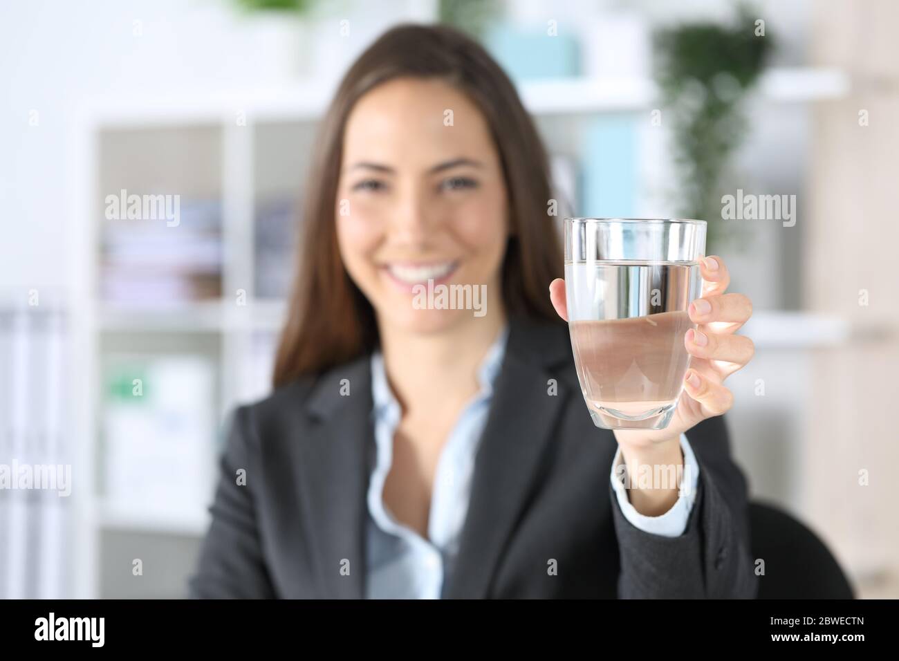 Vue de face d'une femme de direction heureuse montrant un verre d'eau assis sur un bureau Banque D'Images