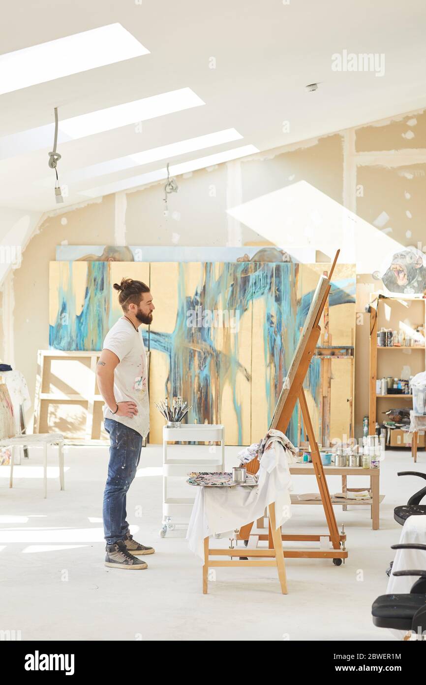 Portrait vertical grand angle d'un artiste contemporain de sexe masculin  regardant la peinture en se tenant debout près d'un chevalet dans un studio  d'art éclairé par le soleil, espace de copie Photo