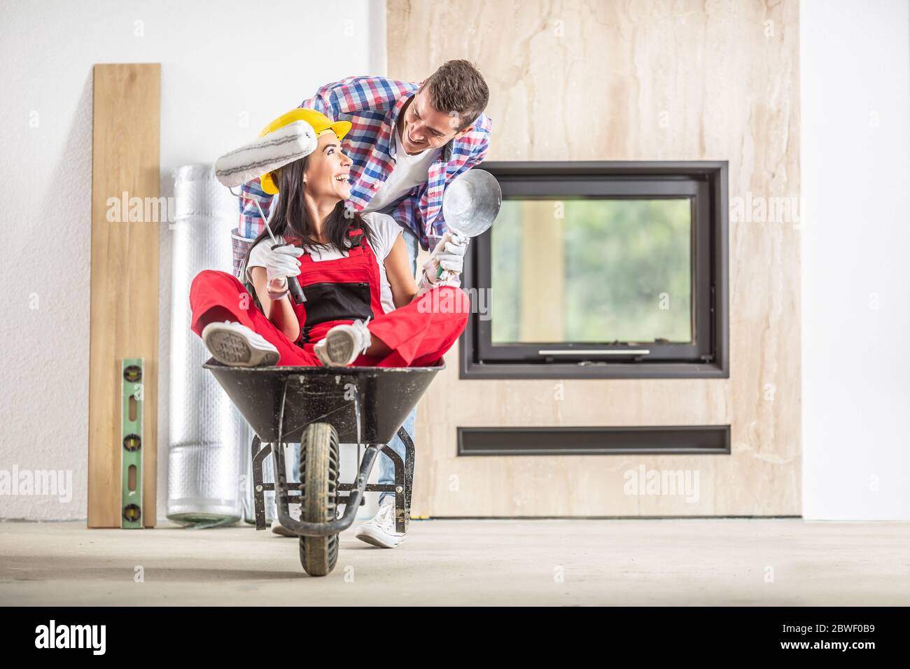 Couple heureux ayant plaisir à monter sur une brouette autour de la maison nouvellement construite, fille dans un casque assis à l'intérieur de la brouette avec des outils dans les mains. Banque D'Images