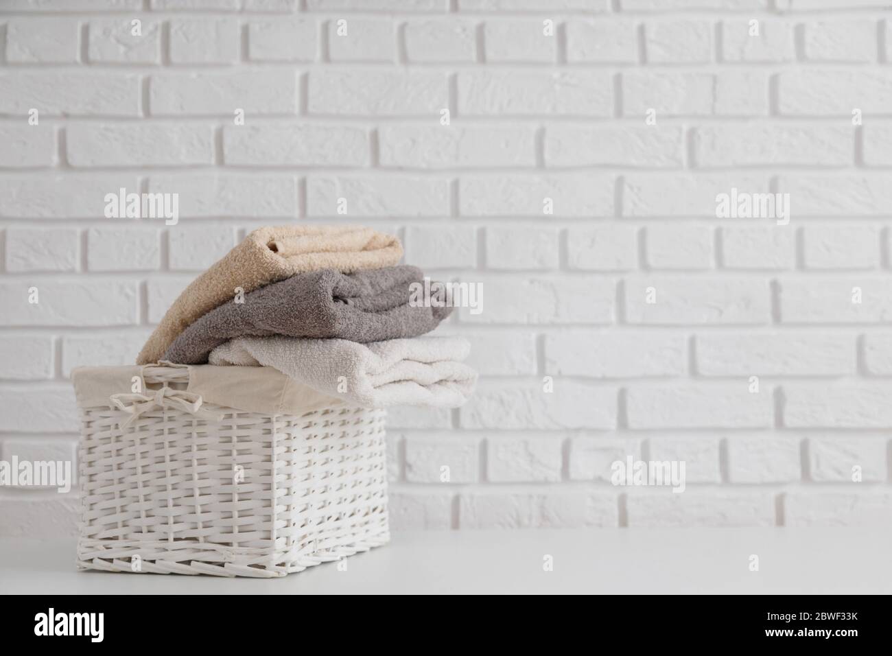 Serviettes fraîches multicolores dans un panier en paille d'osier sur une table blanche avec un mur en brique blanche sur toile de fond. Banque D'Images