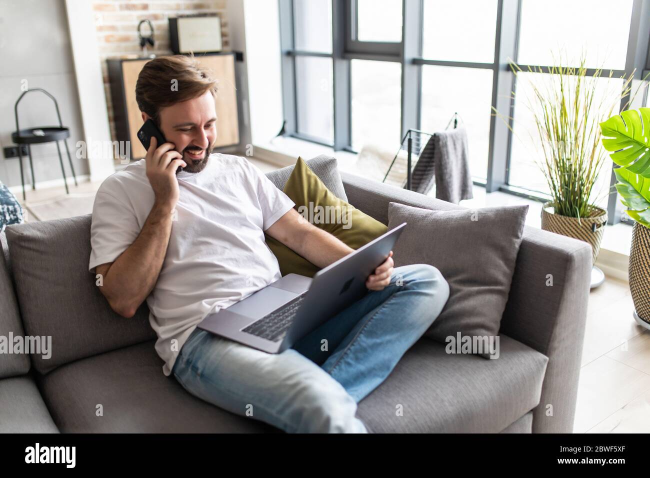 Jeune homme utilisant un ordinateur portable tout en parlant sur le téléphone assis sur le canapé Banque D'Images