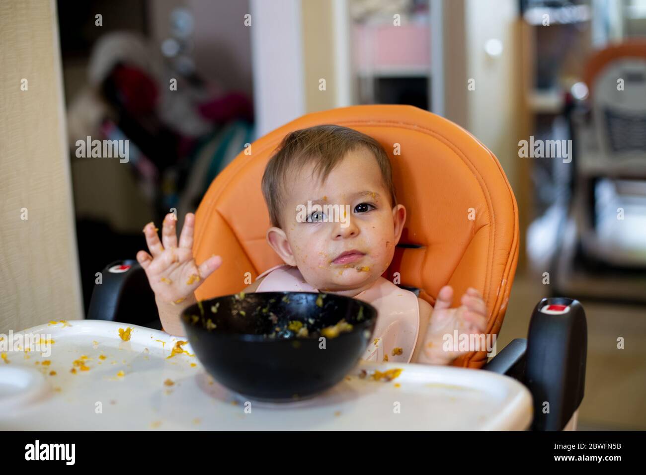 Une Petite Fille Mignonne Dans Un Siege D Enfant Orange Devant Une Assiette De Nourriture Jette Ses Mains Et Regarde L Appareil Photo Tout Autour Dans Des Miettes De Nourriture Gros Plan Photo Stock