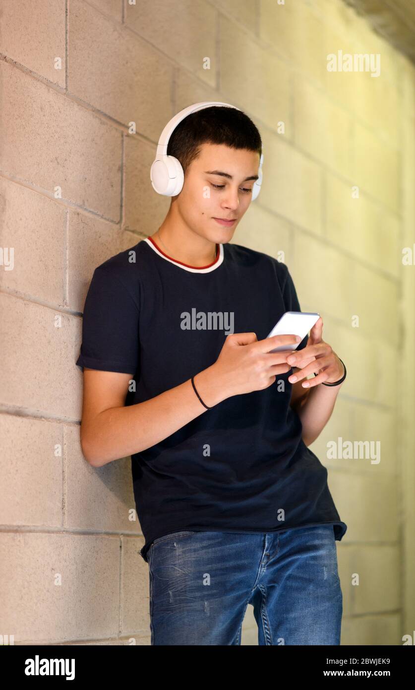 Un jeune adolescent qui écoute de la musique en ville se tenant debout contre un mur en sélectionnant une bande son sur son téléphone mobile pendant qu'il écoute le son Banque D'Images