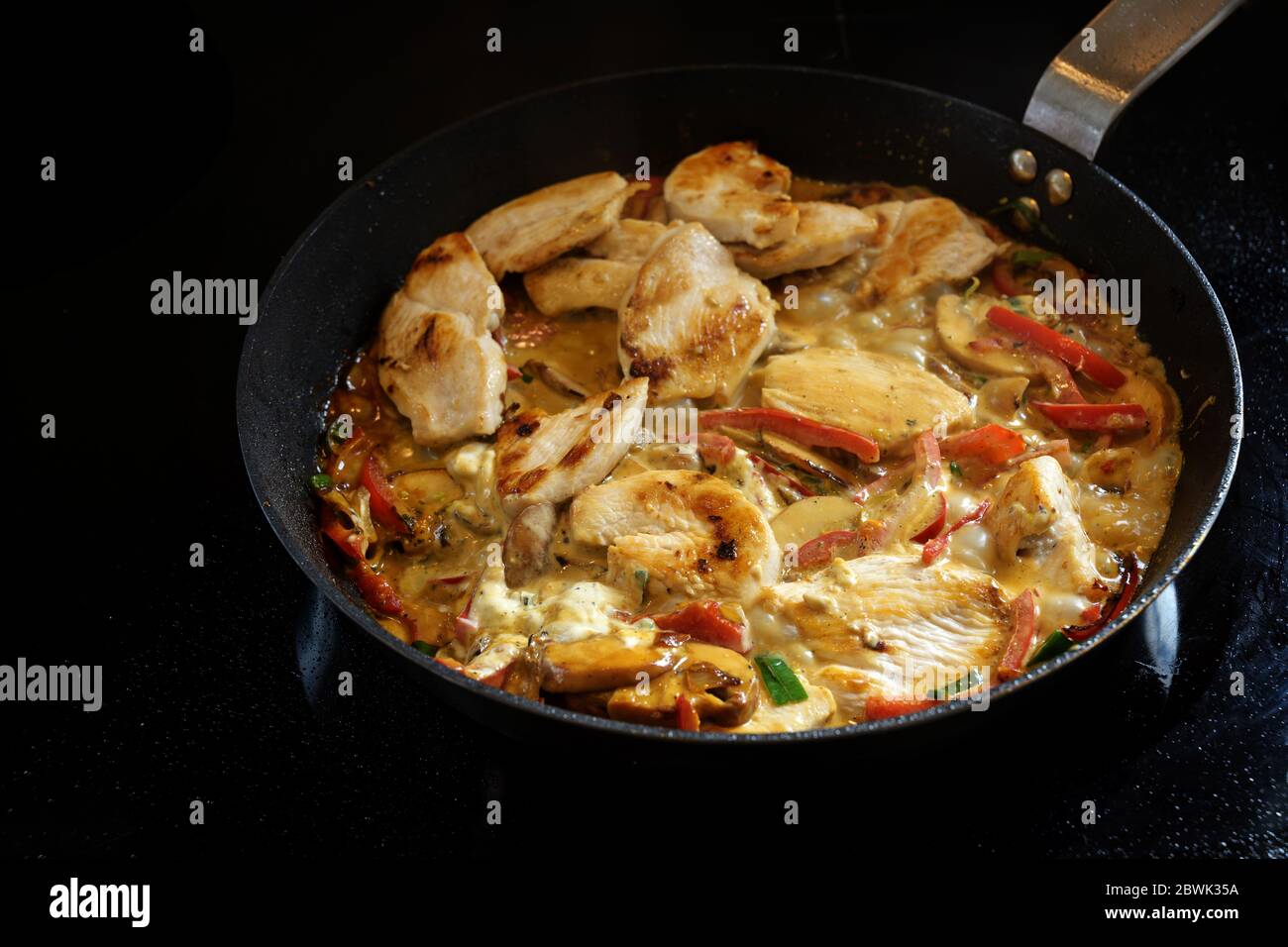 Cuire un ragoût de poulet crémeux avec du poivron et des champignons dans une casserole sur un poêle noir, copier l'espace, le foyer sélectionné, la profondeur de champ étroite Banque D'Images