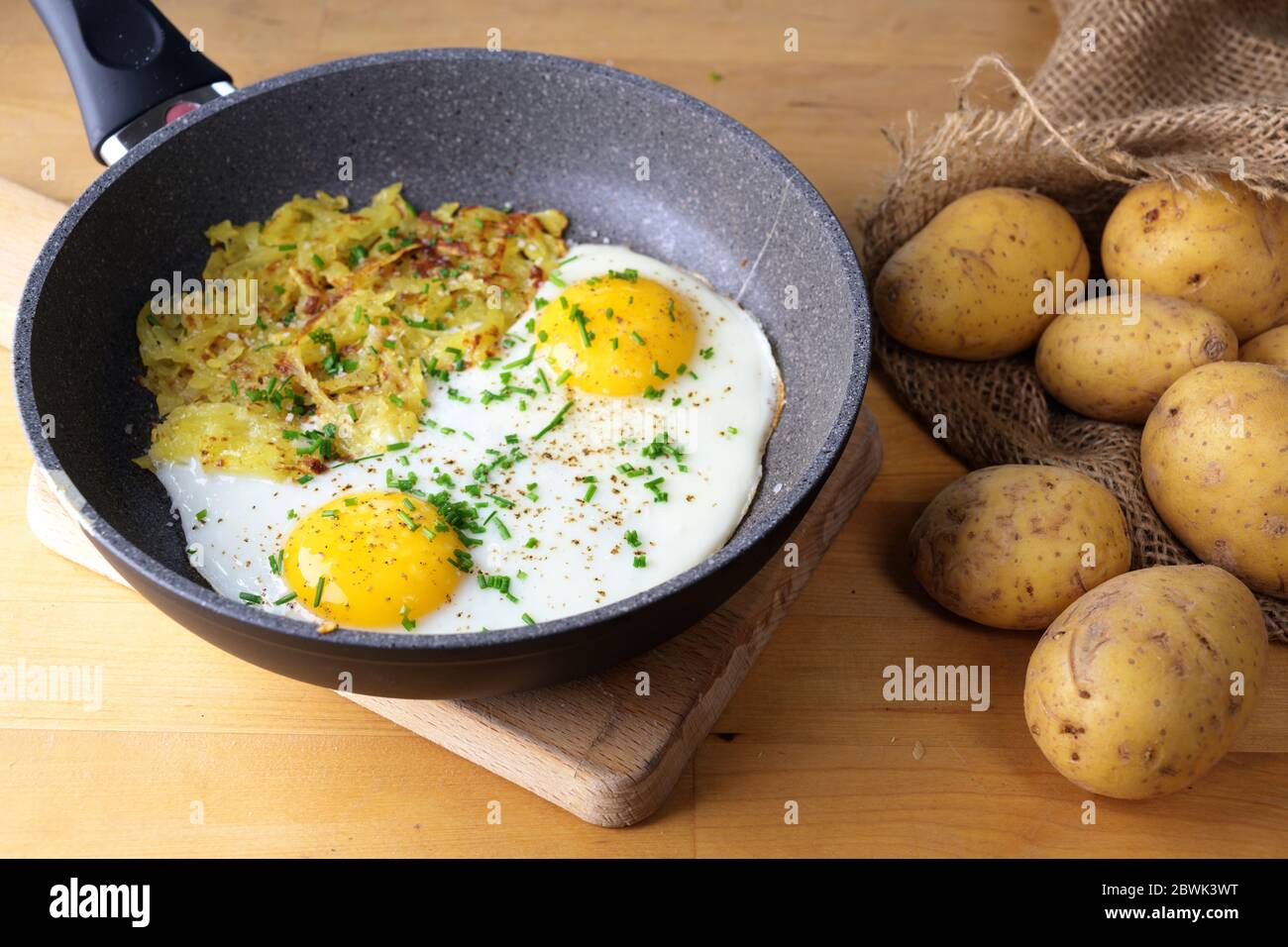 Oeufs frits et roesti suisse dans une poêle sur une table de cuisine en bois avec un sac de pommes de terre, foyer choisi, profondeur de champ étroite Banque D'Images