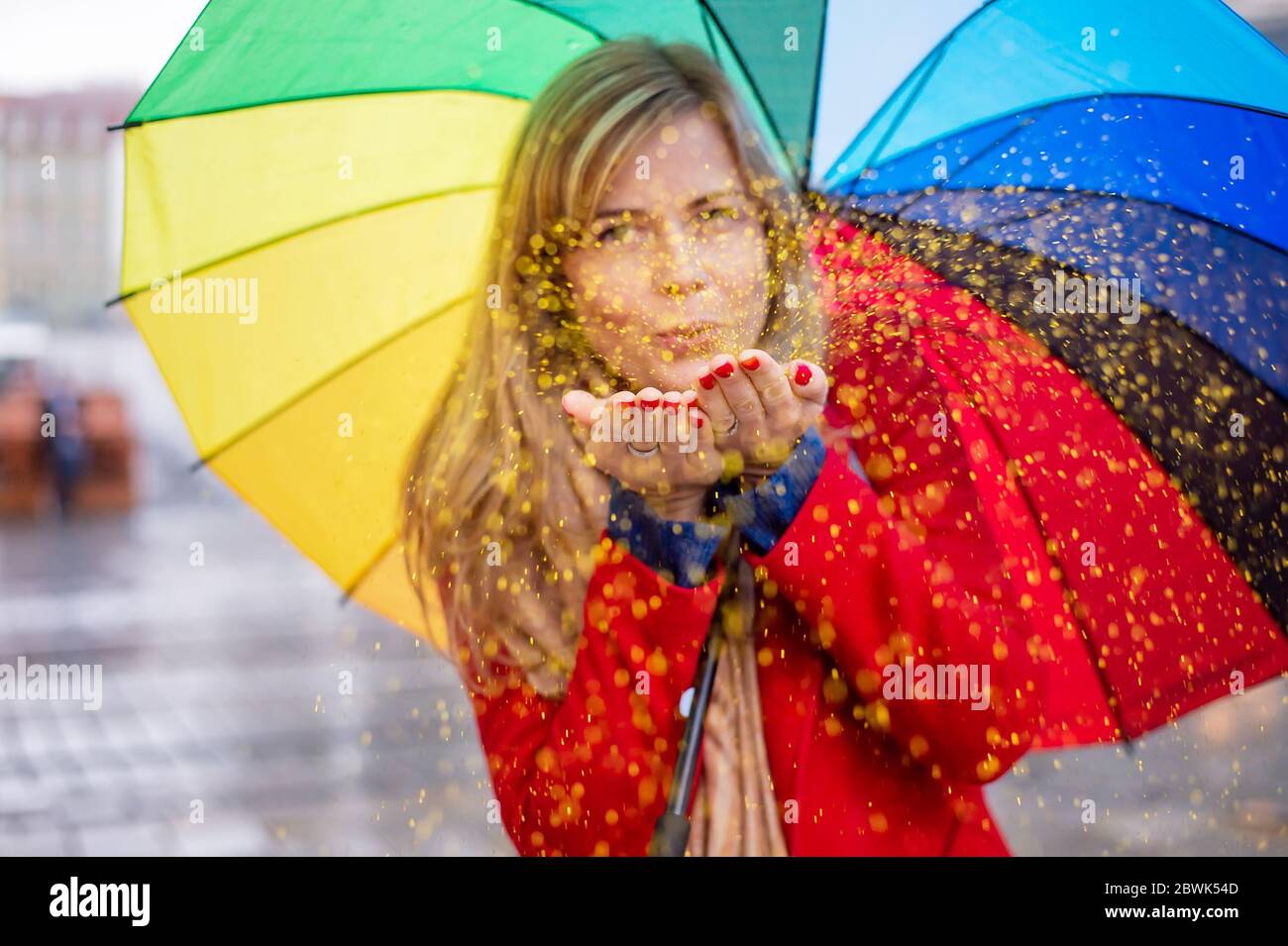 jeune femme blonde tenant un parapluie de couleur arc-en-ciel dans une ambiance festive le jour de pluie. confetti de paillettes soufflantes Banque D'Images