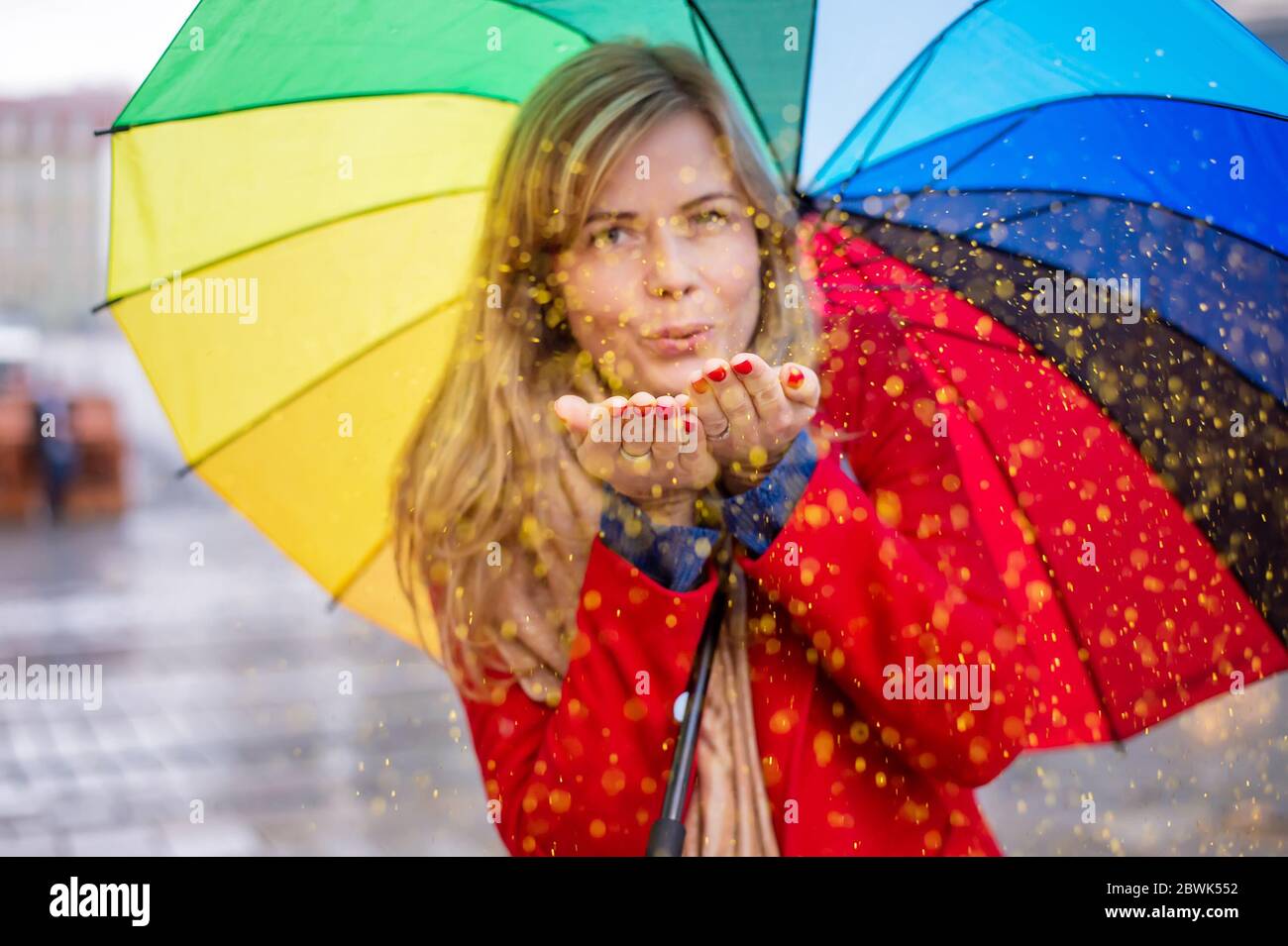 jeune femme blonde tenant un parapluie de couleur arc-en-ciel dans une ambiance festive le jour de pluie. confetti de paillettes soufflantes Banque D'Images