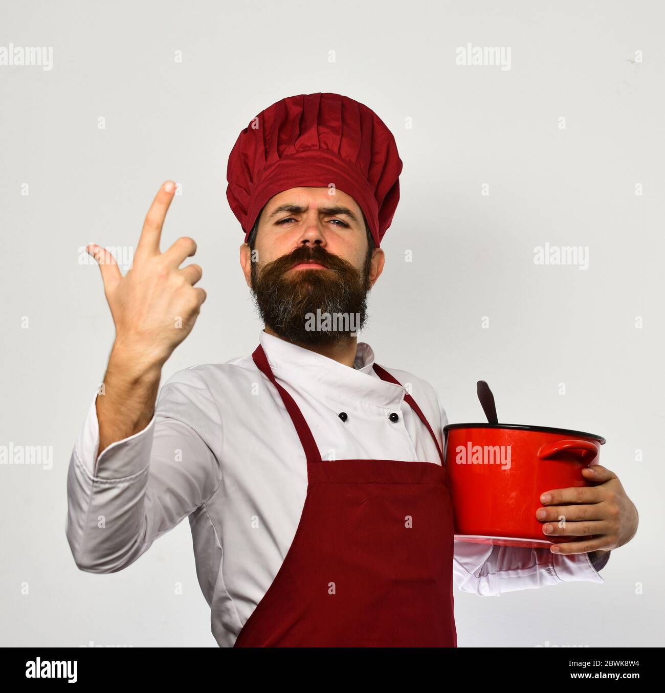 L'homme avec la barbe tient la marmite rouge sur fond blanc. Concept de  ustensiles de cuisine. Cuire avec fierté visage en uniforme bordeaux a  cocotte avec louche. Le chef tient la soupe