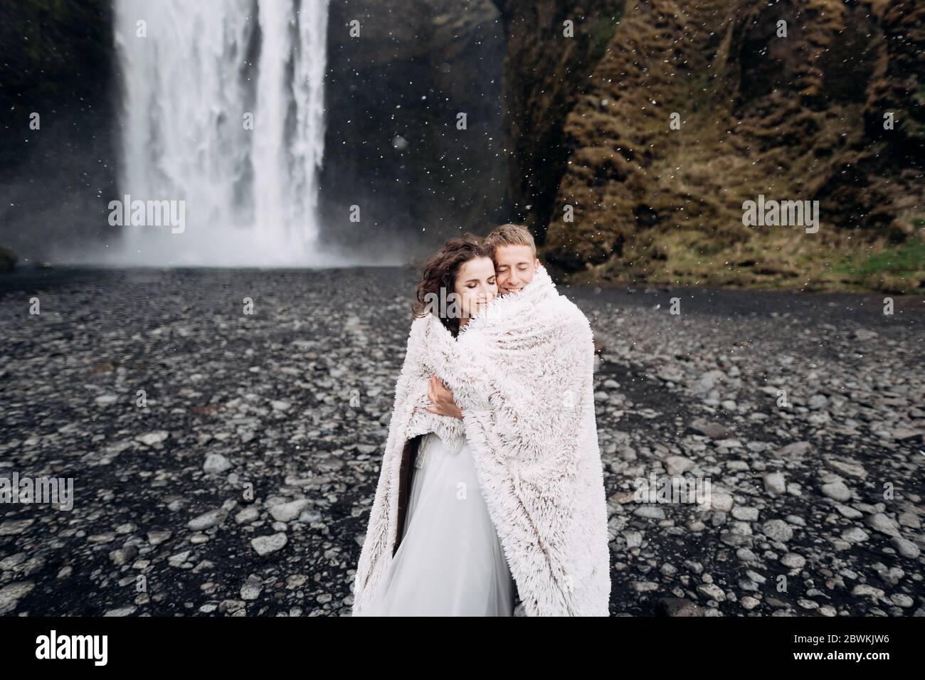 Couple de mariage près de la cascade de Skogafoss. La mariée et le marié recouverts d'une couverture en laine, où embrasser. La neige chute, des flocons de neige tombent. Banque D'Images