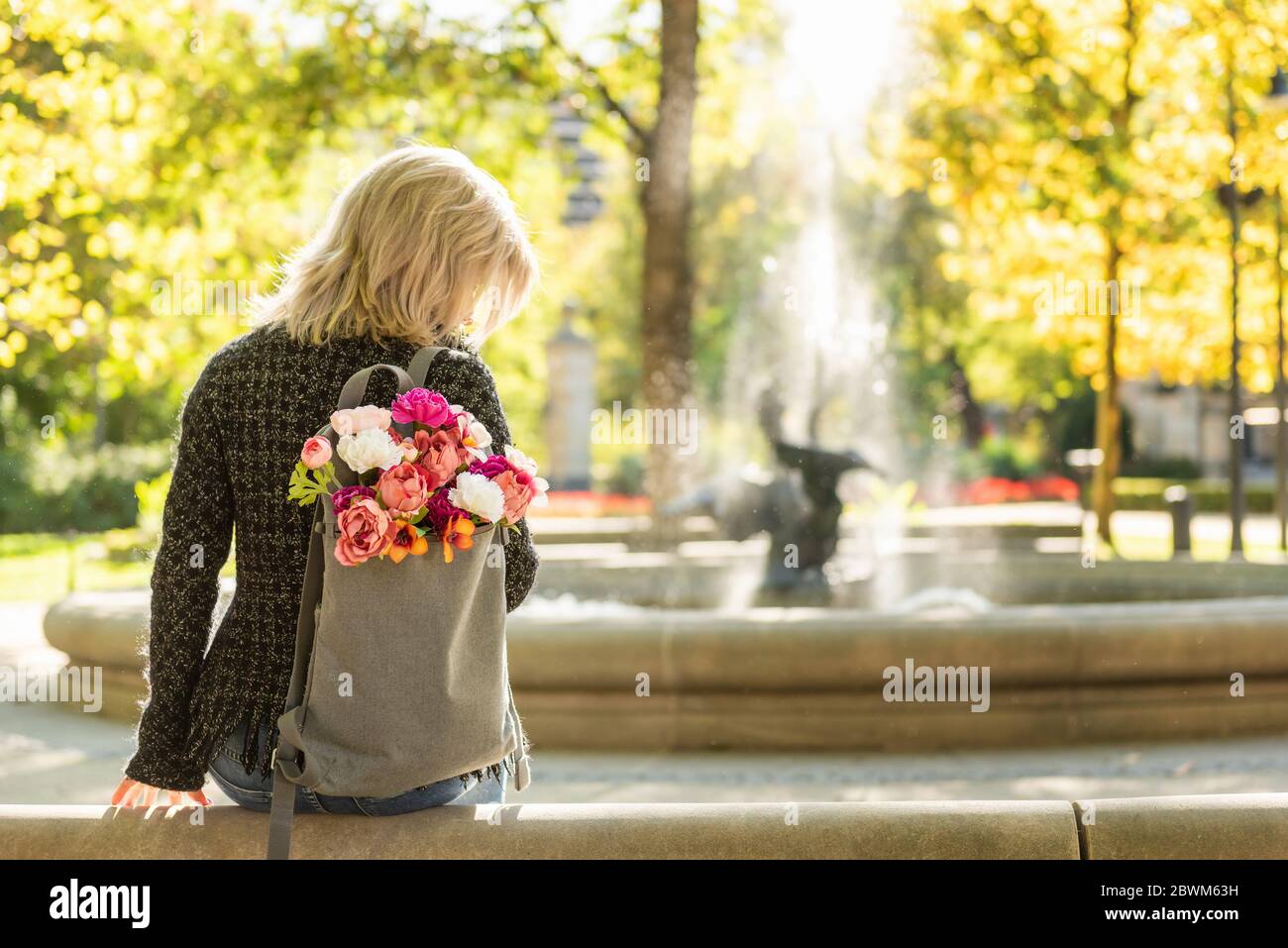 Photo de l'arrière de la jeune femme blonde rêveuse avec bouquet de fleurs dans sac à dos gris dans un parc d'été de printemps Banque D'Images