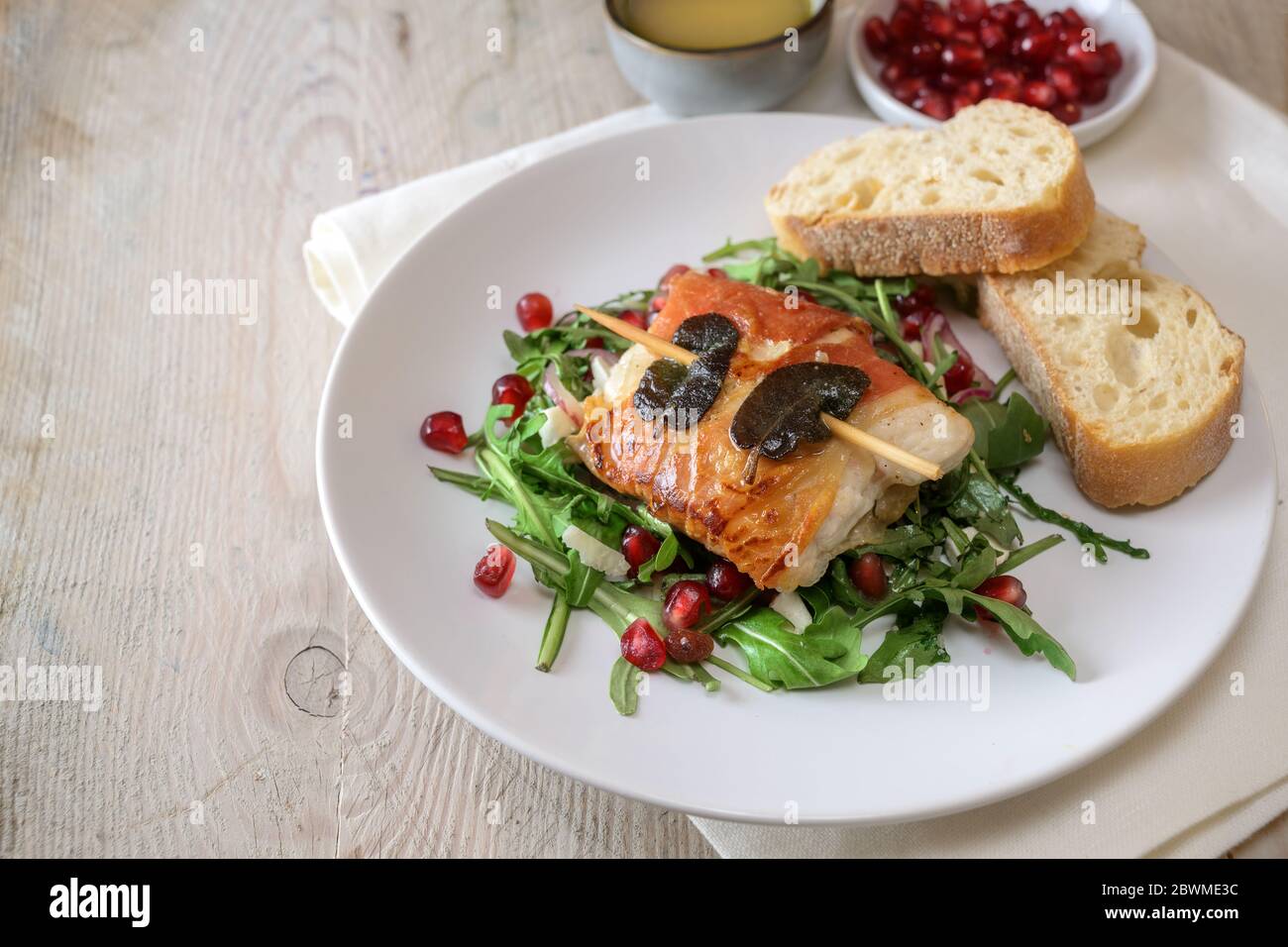 Saltimbocca de poulet rôti enveloppé de jambon prosciutto et de feuilles de sauge, servi avec du pain sur une salade d'arugula sur une assiette blanche, table rustique en bois, copie Banque D'Images