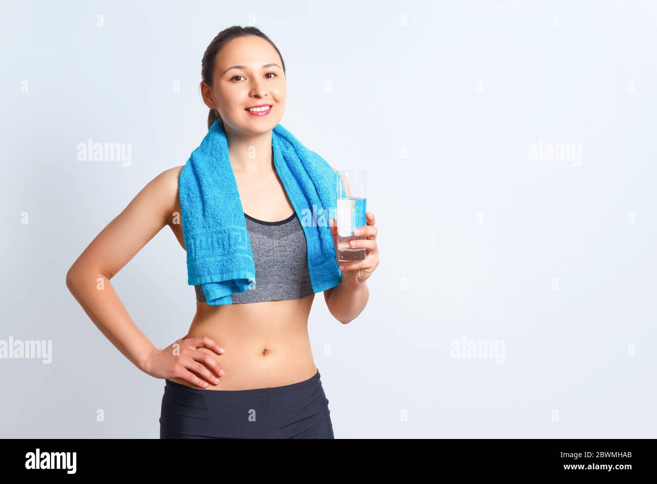Portrait d'une jeune femme de fitness heureuse en vêtements de sport avec une serviette sur ses épaules et un verre d'eau sur fond bleu Banque D'Images