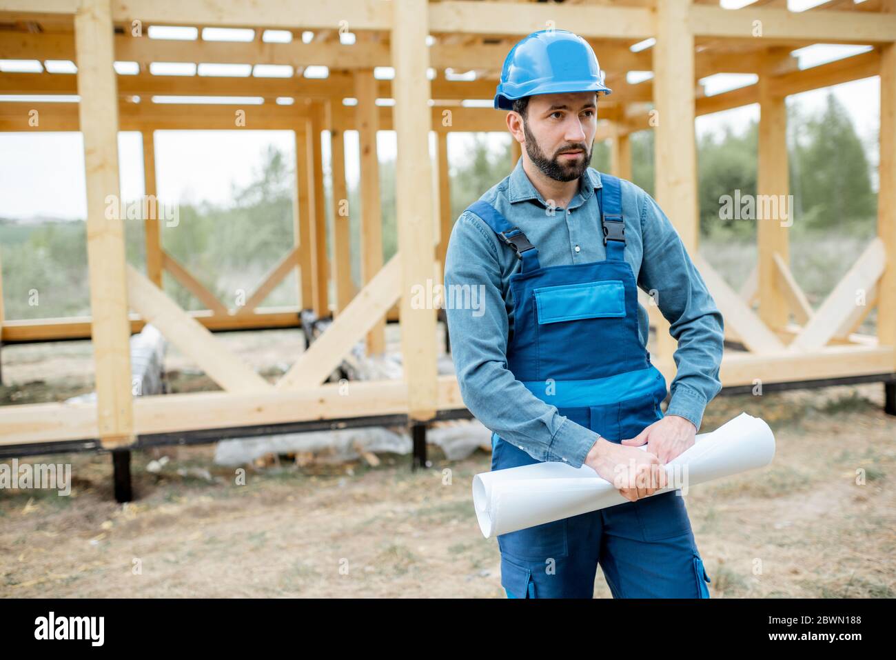 Constructeur en combinaison bleue et chapeau de dur avec des plans sur le chantier de construction, bâtiment en bois cadre maison Banque D'Images