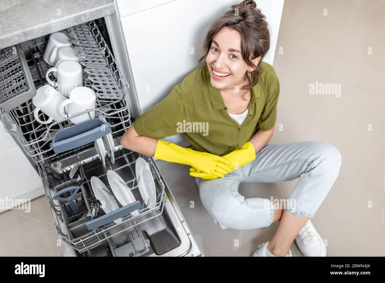 Portrait d'une femme au foyer heureuse assise près du lave-vaisselle avec des plats propres sur la cuisine à la maison. Un travail à domicile facile avec des appareils de cuisine concept Banque D'Images