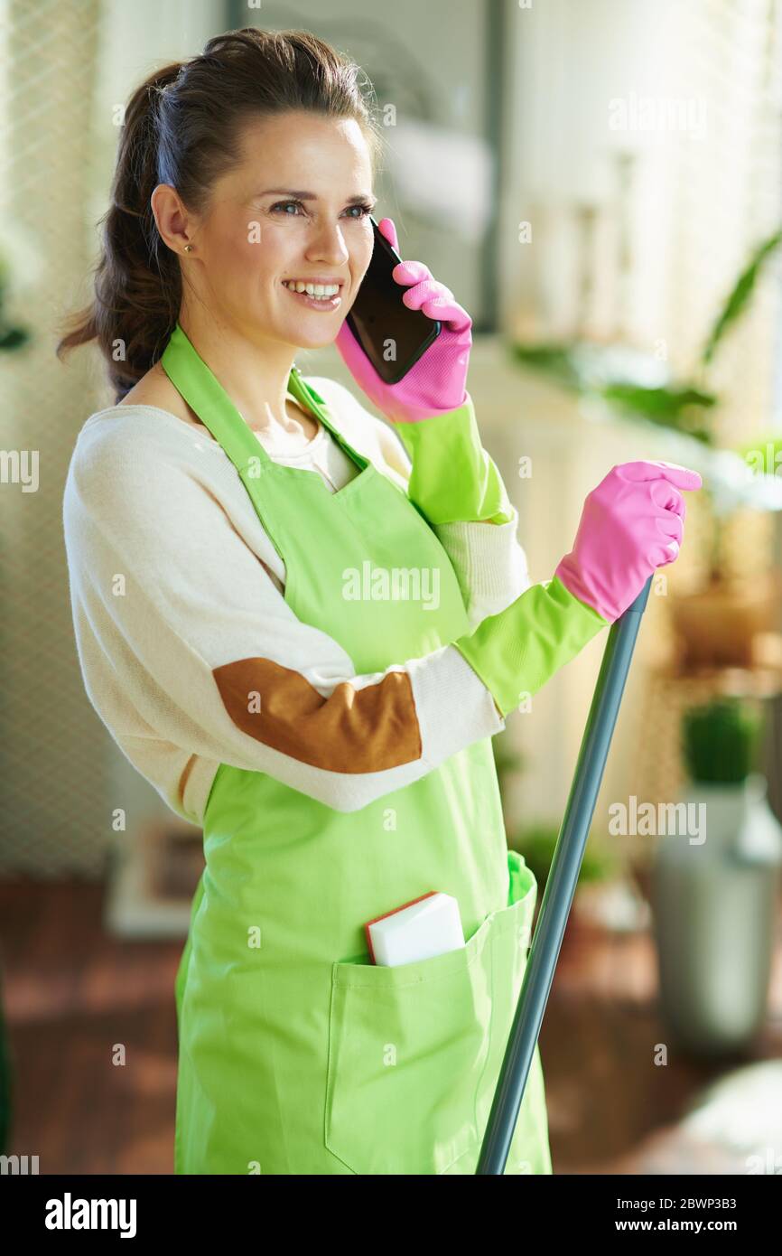 bonne jeune femme au foyer dans un tablier vert et des gants en caoutchouc rose avec une vadrouille parlant sur un téléphone cellulaire dans le salon moderne en journée ensoleillée. Banque D'Images