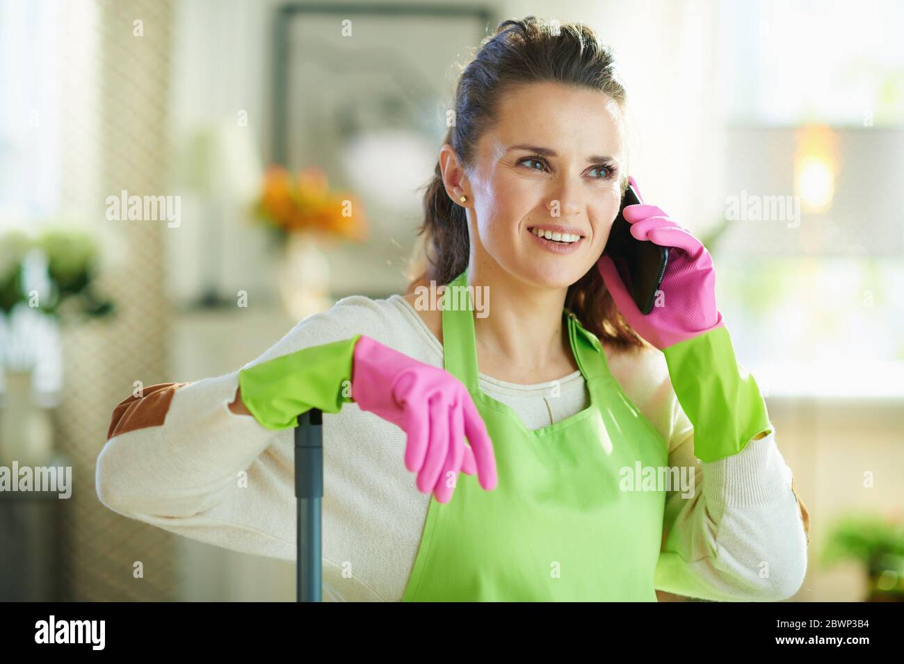 jeune femme souriante avec un tablier vert et des gants en caoutchouc rose avec une vadrouille parlant sur un téléphone cellulaire dans le salon moderne par beau temps. Banque D'Images
