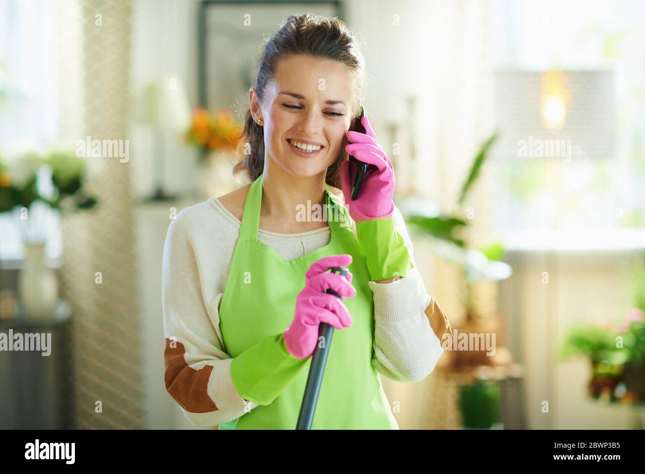 bonne femme moderne d'âge moyen dans un tablier vert et des gants en caoutchouc rose avec une vadrouille parlant sur un téléphone mobile à la maison moderne par temps ensoleillé. Banque D'Images