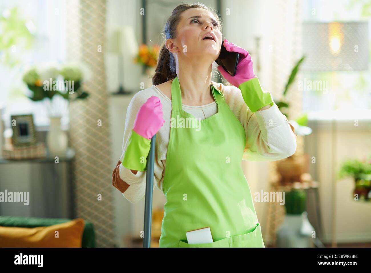 femme moderne fatiguée de 40 ans en tablier vert et gants en caoutchouc rose avec mop utilisant un téléphone mobile dans la maison moderne par temps ensoleillé. Banque D'Images