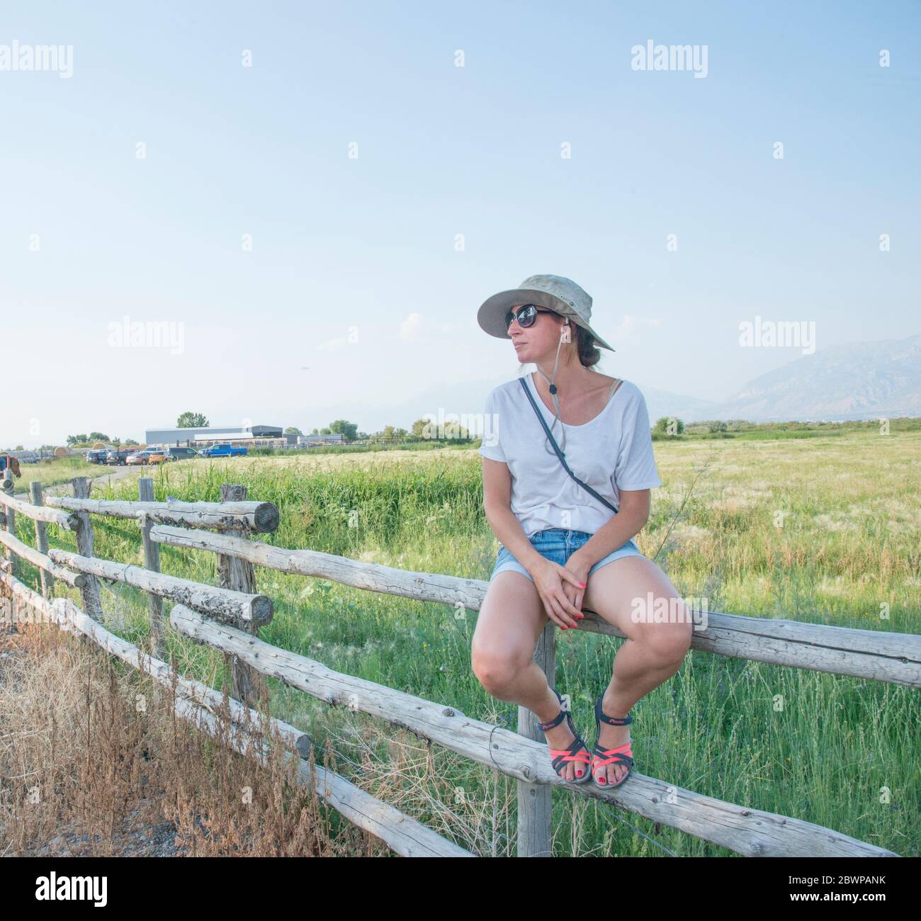 Jeune femme dans un chapeau de soleil assis et posant dessus une clôture en deux parties sur un ranch Banque D'Images