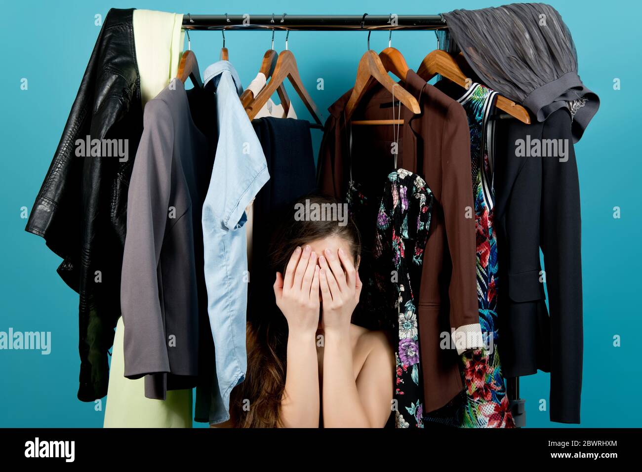 La fille couvre son visage, perdu dans le choix des vêtements sur le fond des cintres avec des vêtements, fond bleu Banque D'Images