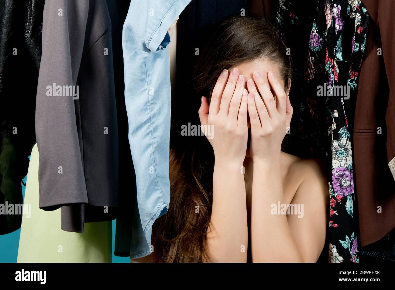 La fille couvre son visage, perdu dans le choix des vêtements sur le fond des cintres avec des vêtements, fond bleu Banque D'Images