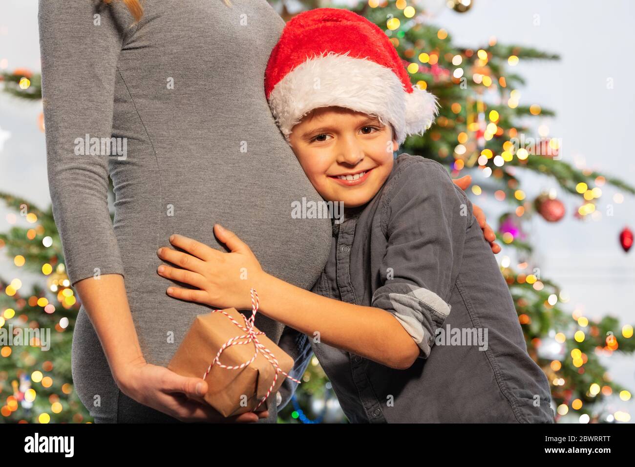 Portrait d'un petit garçon portant un chapeau de père Noël câlin le ventre de la mère tenant présent debout près de l'arbre du nouvel an Banque D'Images