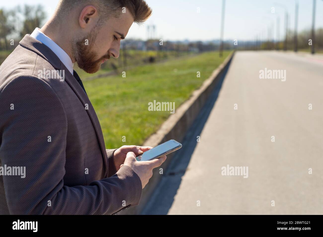 Utilisation du téléphone portable à l'extérieur - avec trafic de ville déféoutilisé. Portrait d'un jeune homme barbu de vingt-cinq ans, utilisant un smartphone, se tient à la CA Banque D'Images