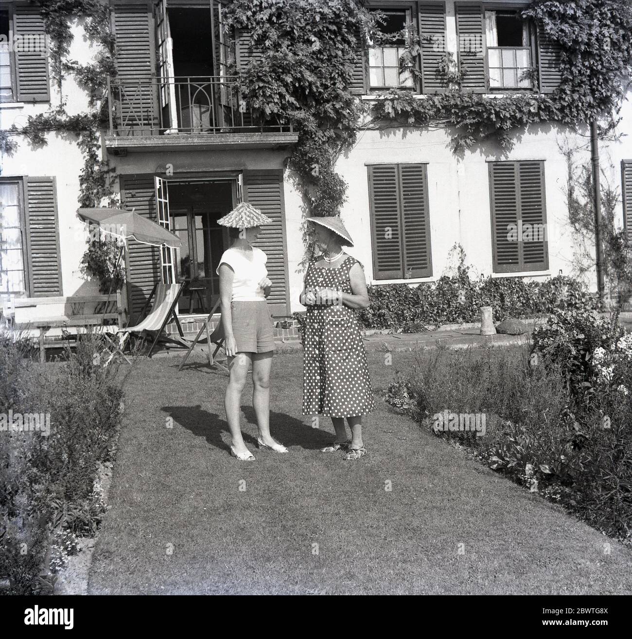 Années 1960, historique, l'été et deux dames debout sur la pelouse à l'extérieur d'une maison continentale avec des volets de fenêtre ayant une conversation et portant des chapeaux de soleil de style chinois 'Coolie'. Banque D'Images