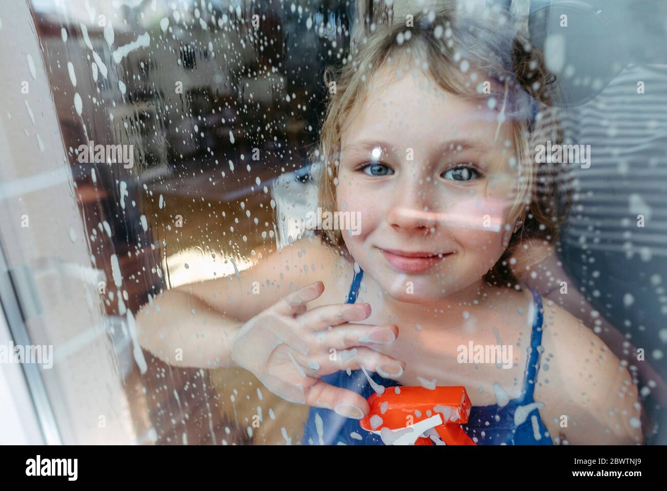 Portrait de fille blonde souriante avec flacon vaporisateur regardant par fenêtre humide Banque D'Images