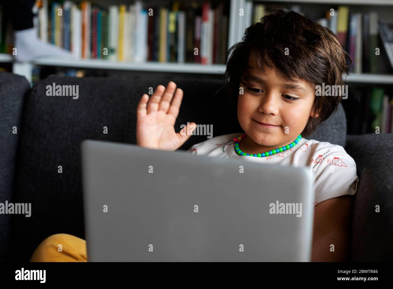 Un adorable garçon agite tout en utilisant un ordinateur portable pour passer des appels vidéo dans le salon Banque D'Images