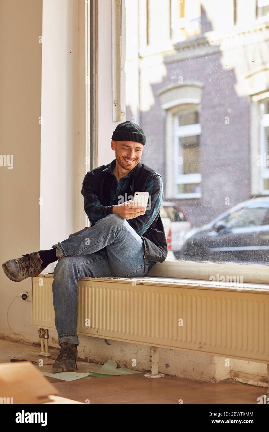 Homme en train de rénover l'atelier, assis sur un rebord de fenêtre, à l'aide d'un smartphone Banque D'Images