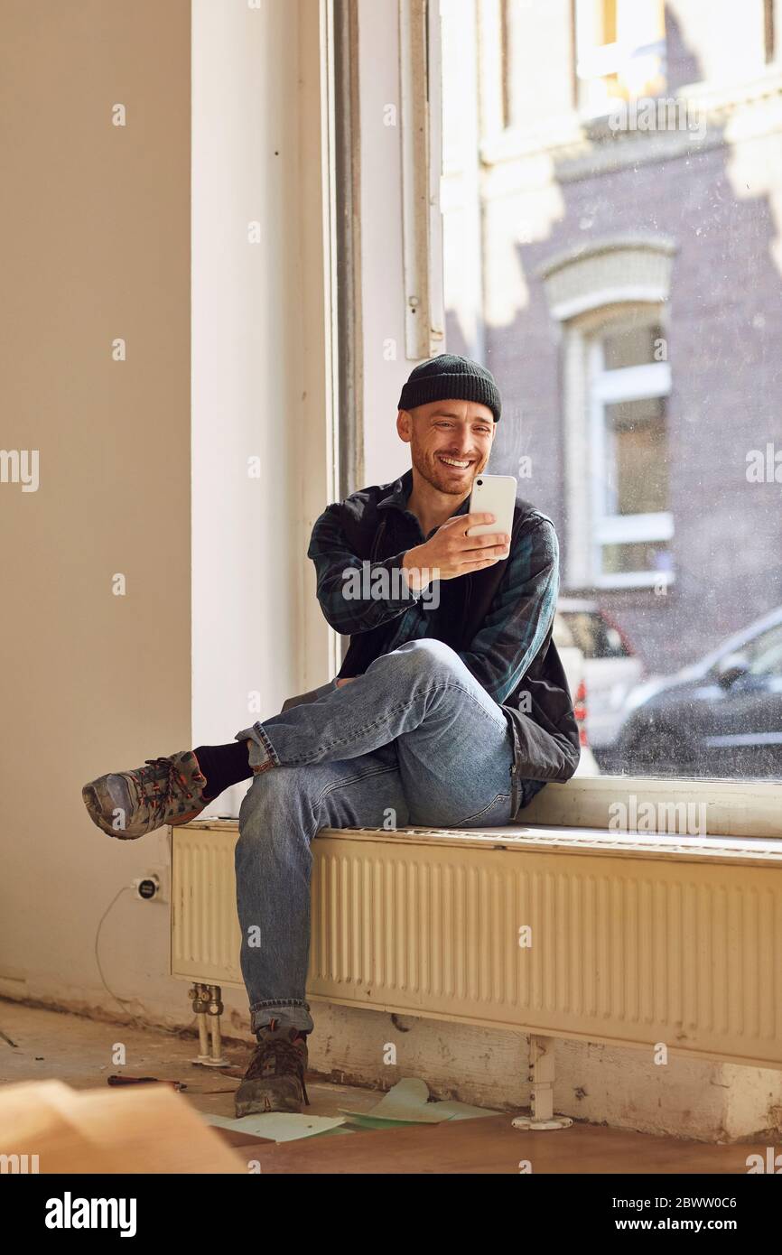Homme en train de rénover l'atelier, assis sur un rebord de fenêtre, à l'aide d'un smartphone Banque D'Images