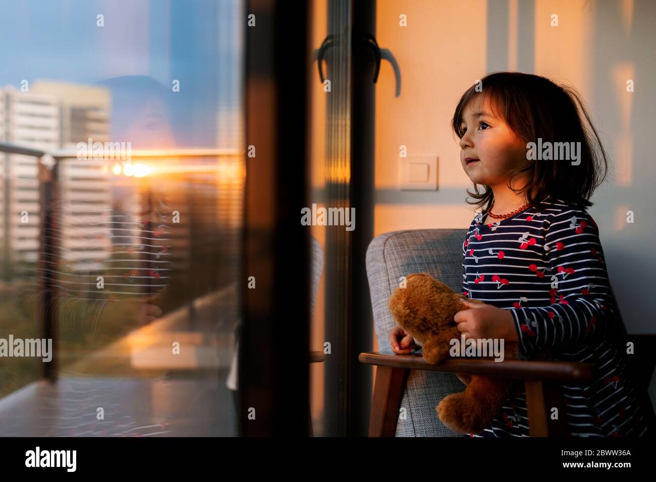 Portrait d'une petite fille avec un ours en peluche, vue par la fenêtre au coucher du soleil Banque D'Images