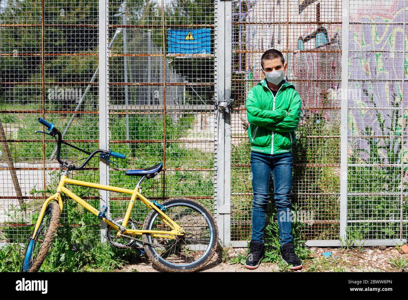 Portrait d'un pré-adolescent portant un masque facial debout avec les bras croisés à vélo contre la clôture Banque D'Images