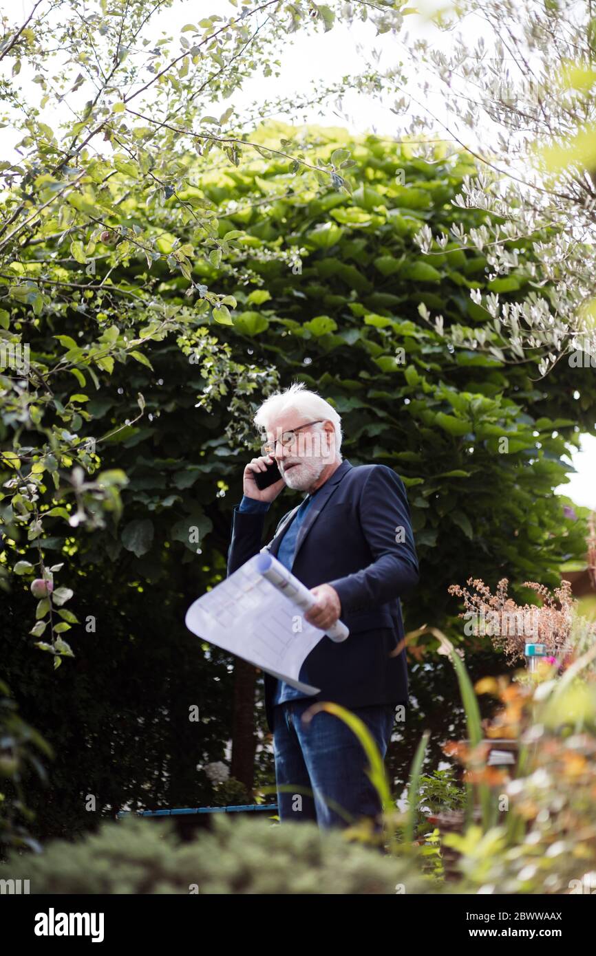 Homme senior au téléphone debout dans le jardin avec plan de construction Banque D'Images