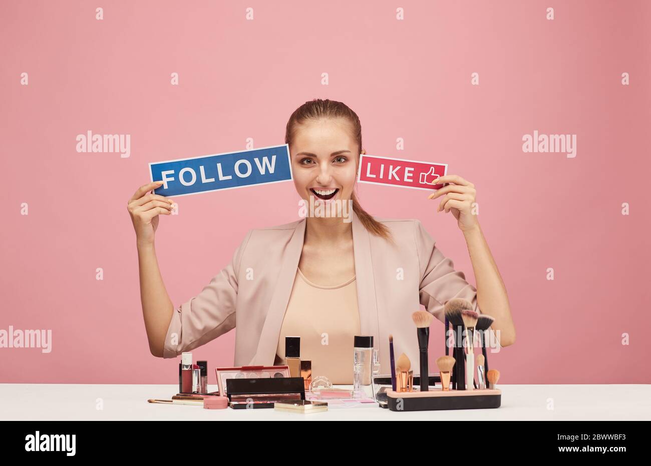 Portrait de la beauté excitée blogueur assis à la table avec des cosmétiques et souriant à l'appareil photo sur fond rose Banque D'Images