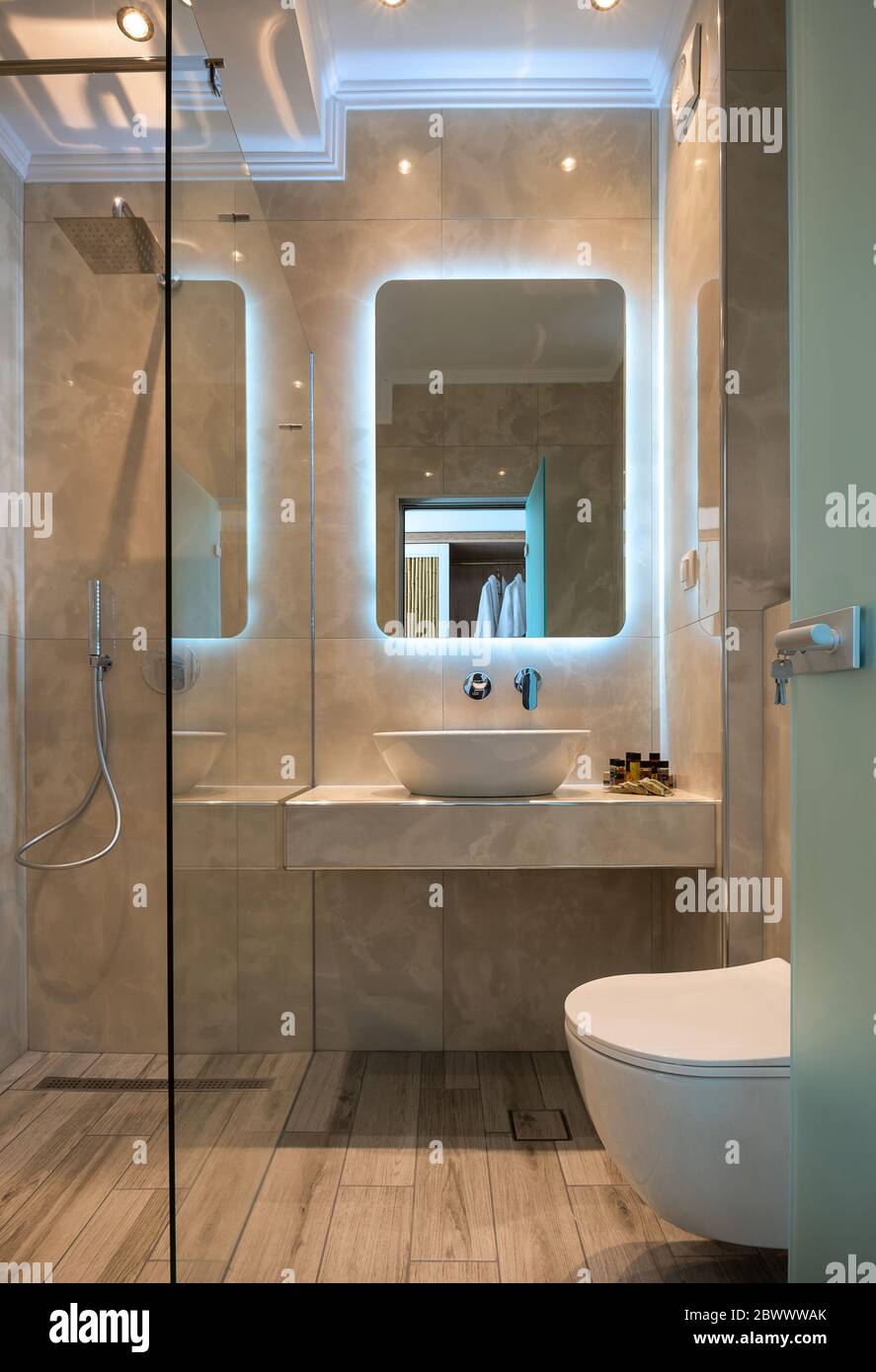 Vue sur la façade de la salle de bains moderne en marbre beige avec lavabo, miroir de lumière, cabine de douche en verre, toilettes murales, parquet Banque D'Images