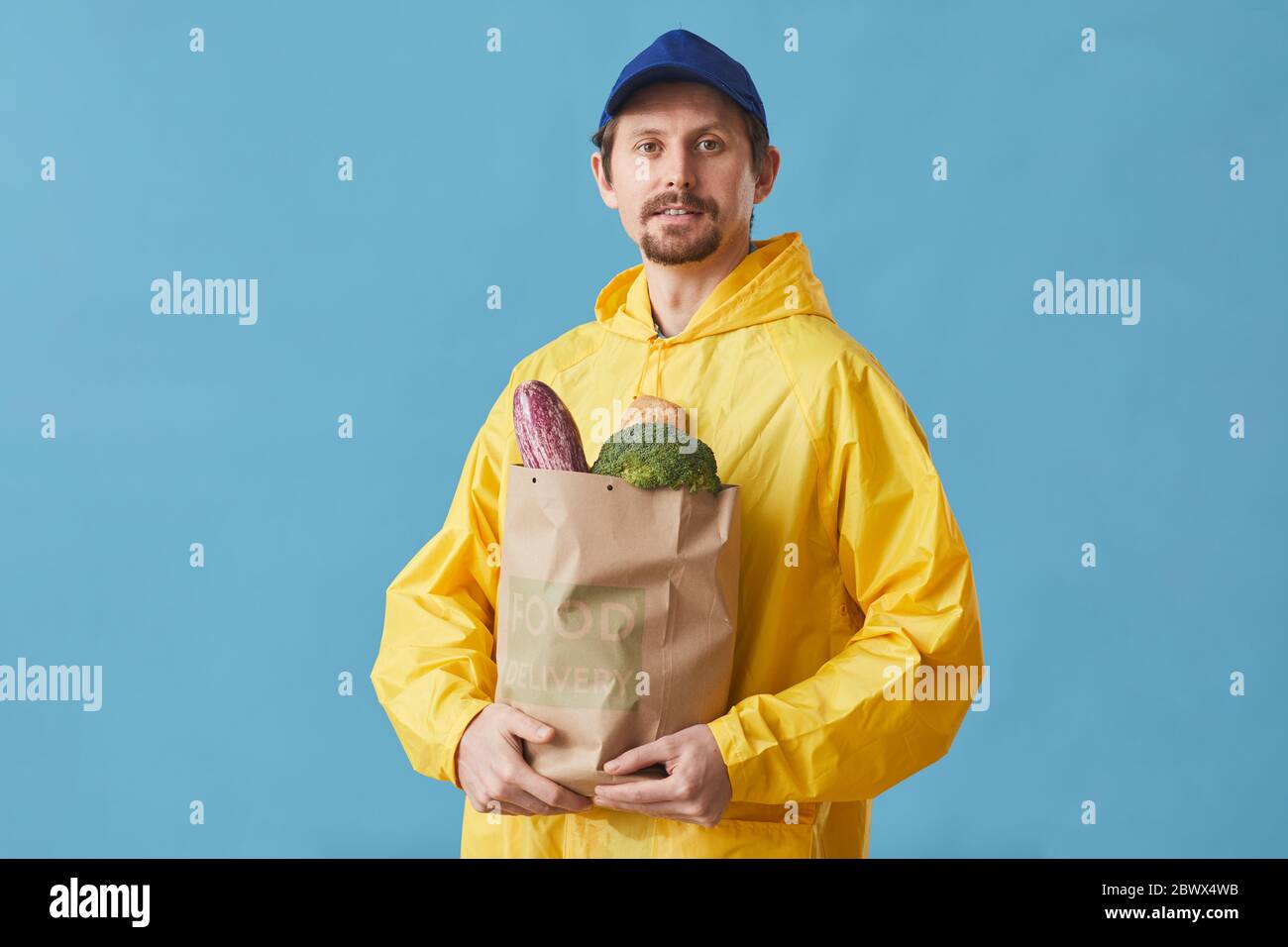 Portrait d'un jeune homme en uniforme tenant un sac en papier avec des produits sur fond bleu, il livrant de la nourriture Banque D'Images