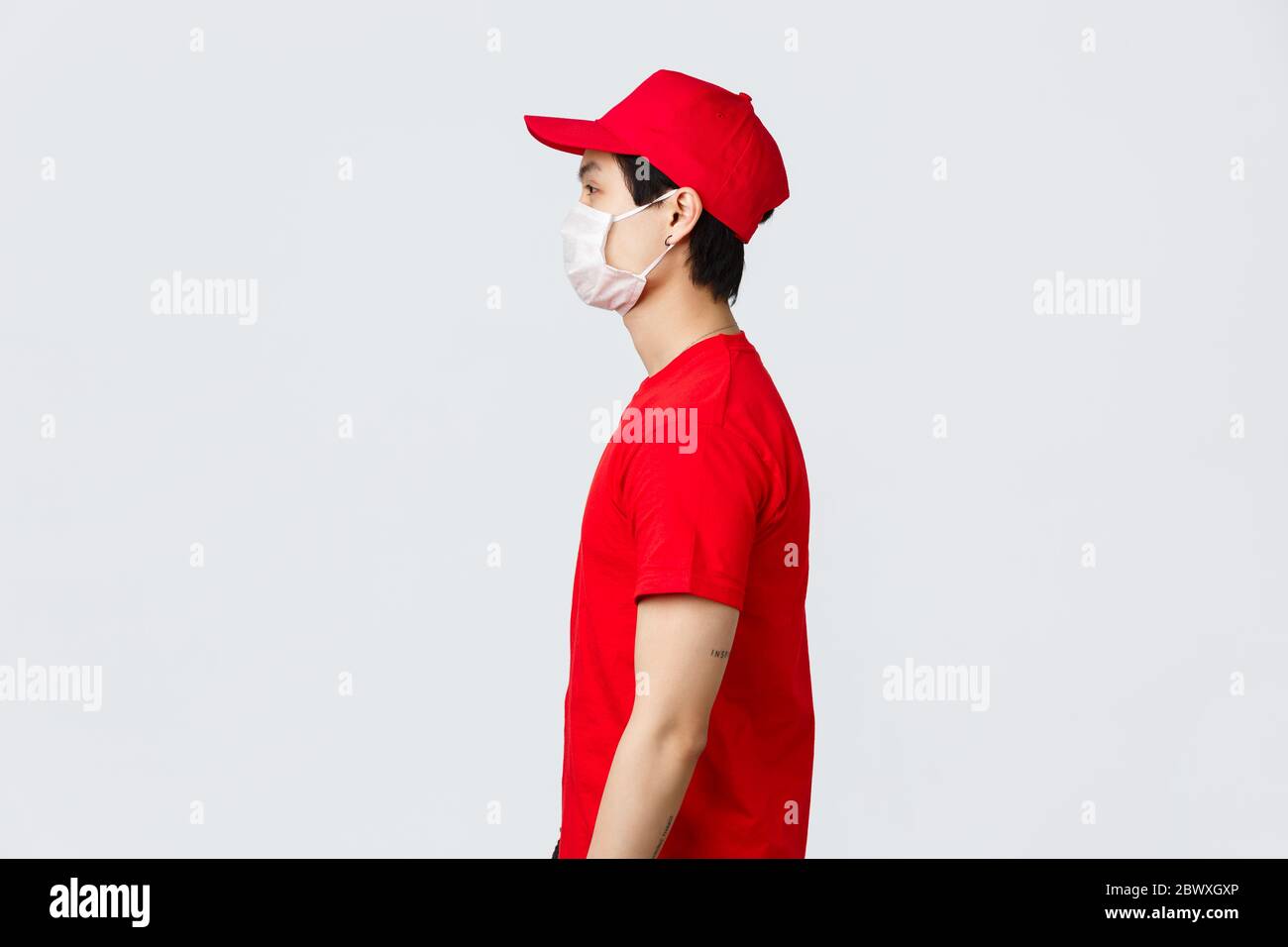 Photo de profil d'un homme de livraison asiatique en casquette rouge et  t-shirt, portant un masque médical de protection, regardant à gauche,  attendant à la porte pour le ramassage du client Photo
