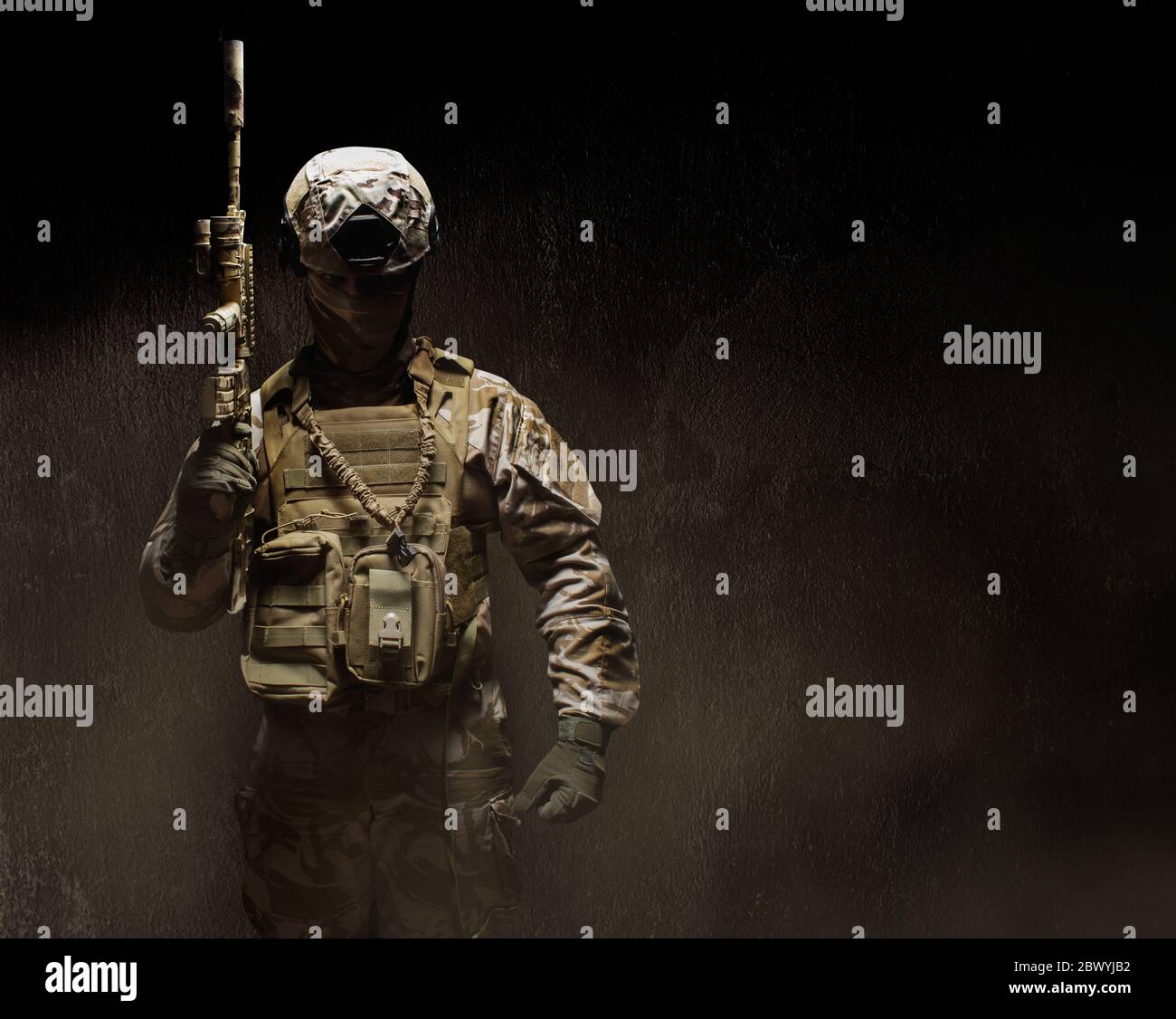 Photo d'un soldat de camouflage du désert entièrement équipé dans un masque, un casque, une armure et des gants debout avec un fusil sur fond de béton brumeux foncé Banque D'Images