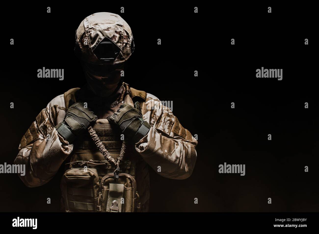 Photo d'un soldat de camouflage du désert entièrement équipé, avec masque, casque, armure et gants, debout sur fond noir et brouillard. Banque D'Images