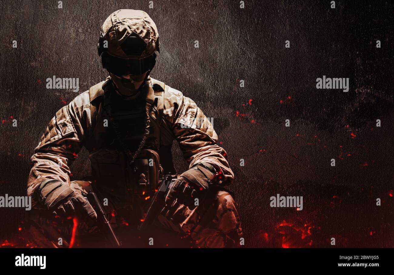 Photo d'un soldat du désert entièrement équipé dans une armure camoufée assise avec des armes à feu et des cendres volantes sur fond de béton grundy. Banque D'Images