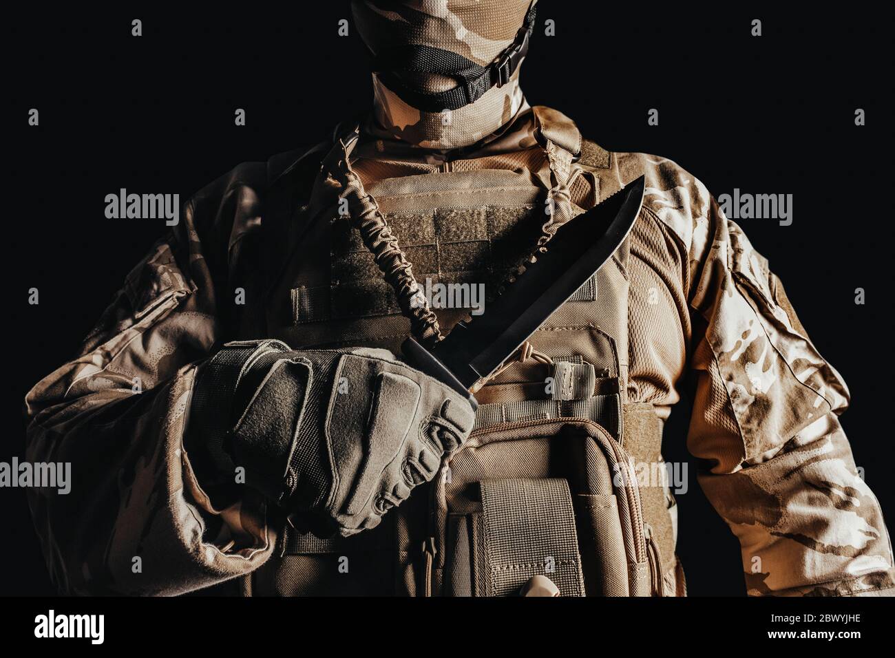 Photo d'un soldat entièrement équipé dans une armure camoufée et des gants tenant un couteau militaire sur fond noir. Banque D'Images