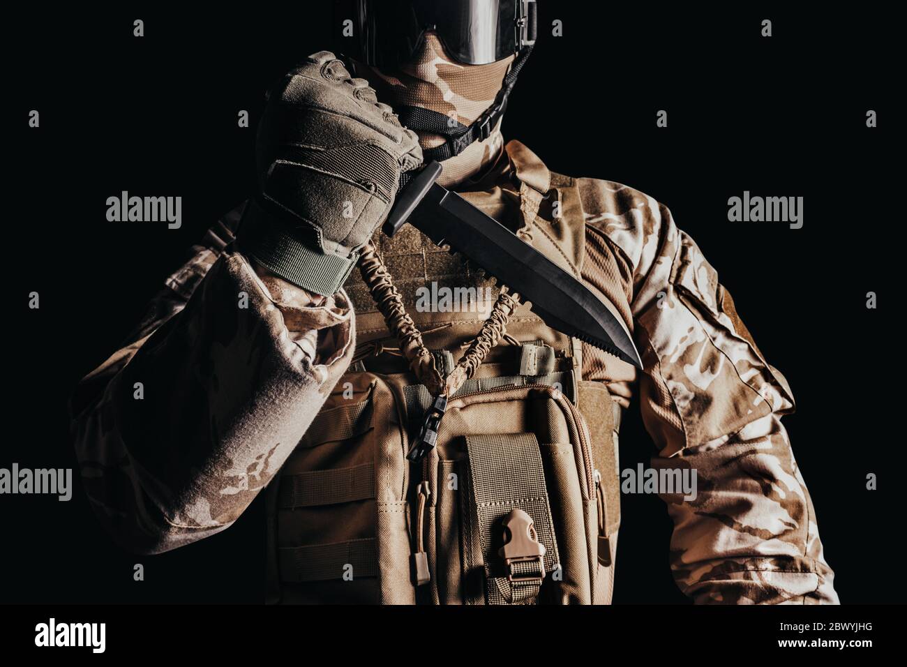 Photo d'un soldat entièrement équipé dans une armure camoufée et des gants tenant un couteau militaire à prise inversée sur fond noir. Banque D'Images