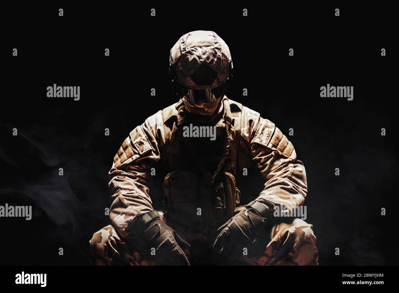 Photo d'un soldat du désert entièrement équipé dans une armure camouflée, un casque et des gants assis sur un fond sombre et brumeux. Banque D'Images