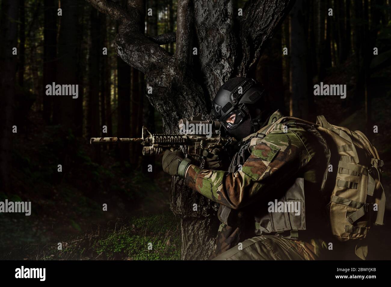 Photo d'un soldat entièrement équipé en uniforme de camouflage et sac à dos assis et visant le fusil près de l'arbre sur fond de forêt sombre. Banque D'Images
