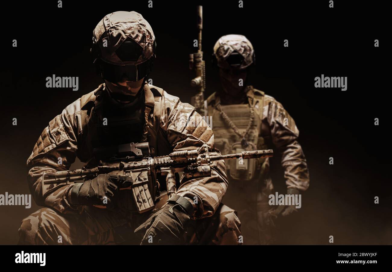 Photo d'un soldat du désert entièrement équipé dans une armure camoufée assise et debout avec un fusil sur fond sombre et brumeux. Banque D'Images