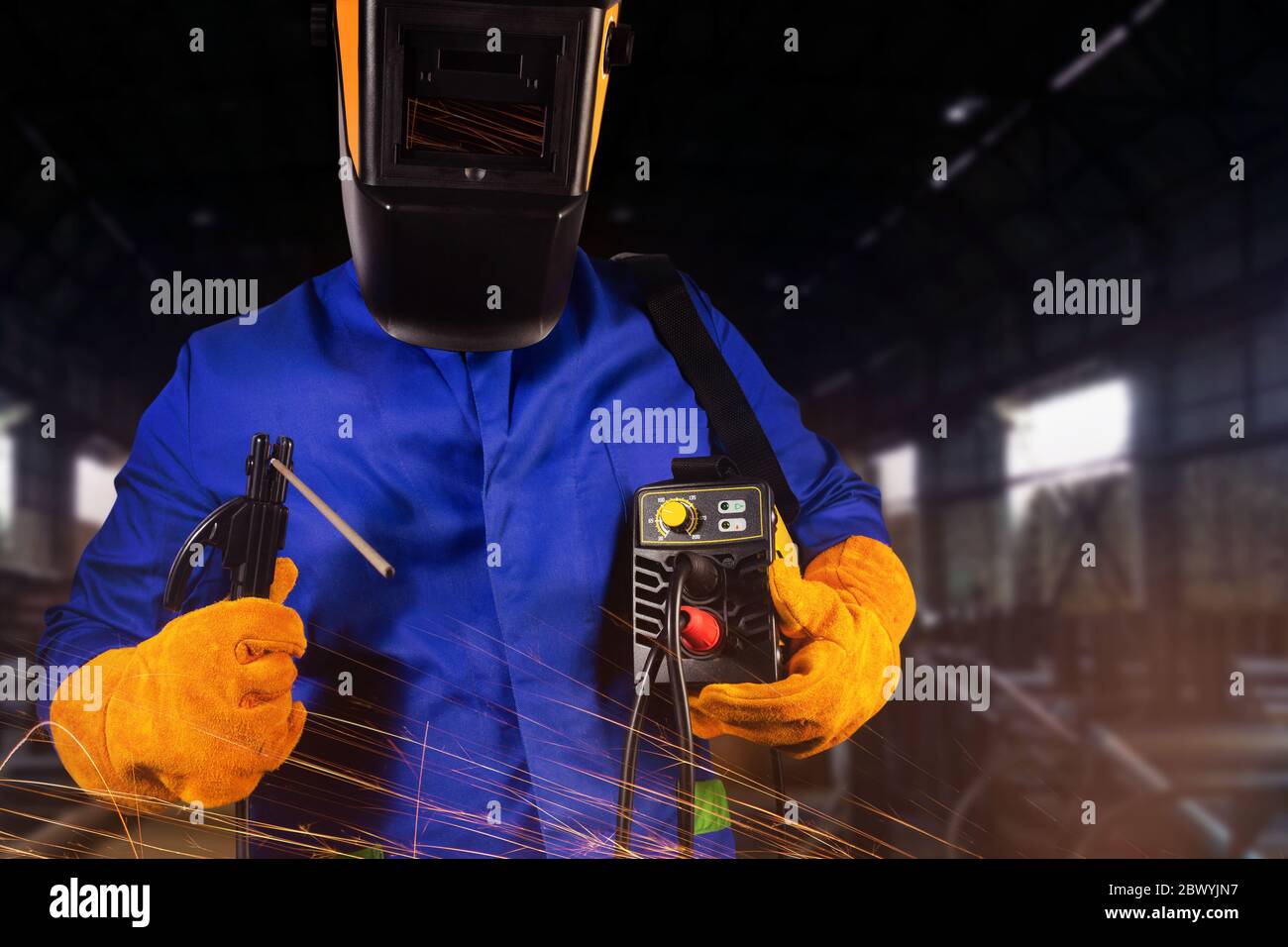 Photo d'un soudeur en uniforme, casque et gants tenant l'inverseur et les électrodes sur un fond industriel. Banque D'Images